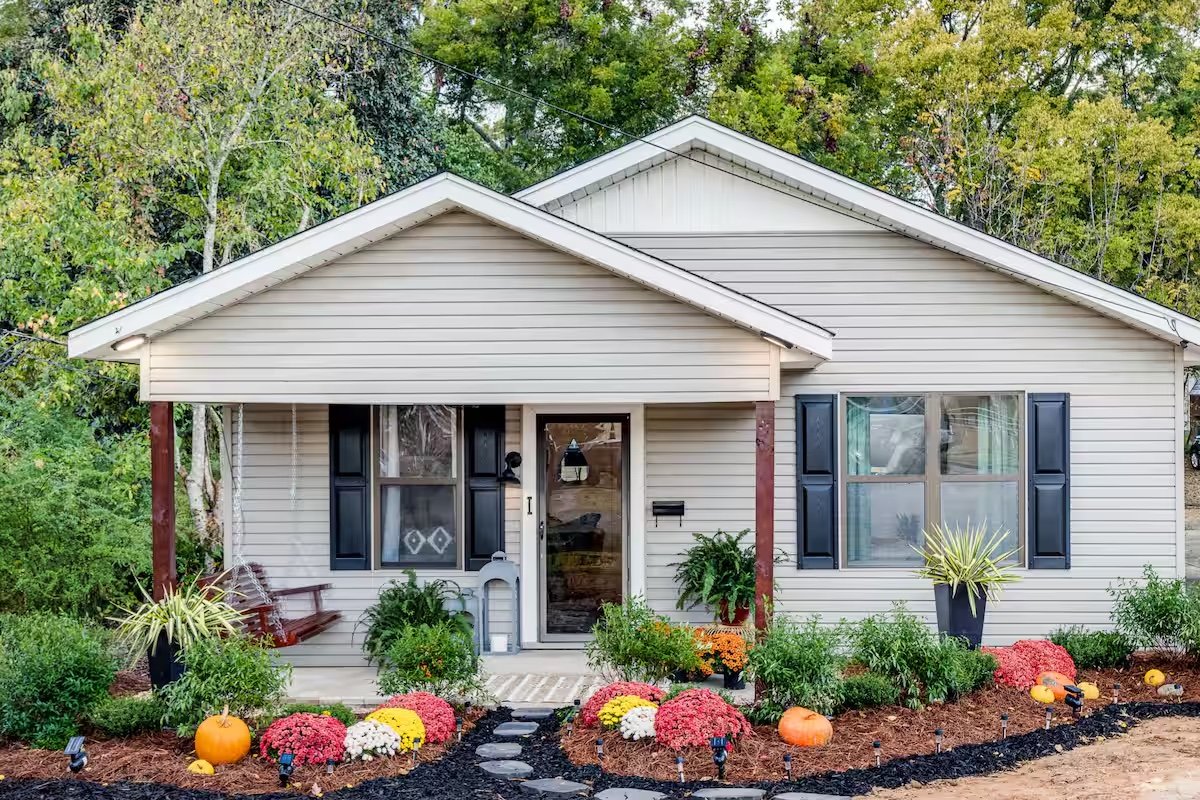 A gray bungalow with black shutters, a porch swing and fall flower beds