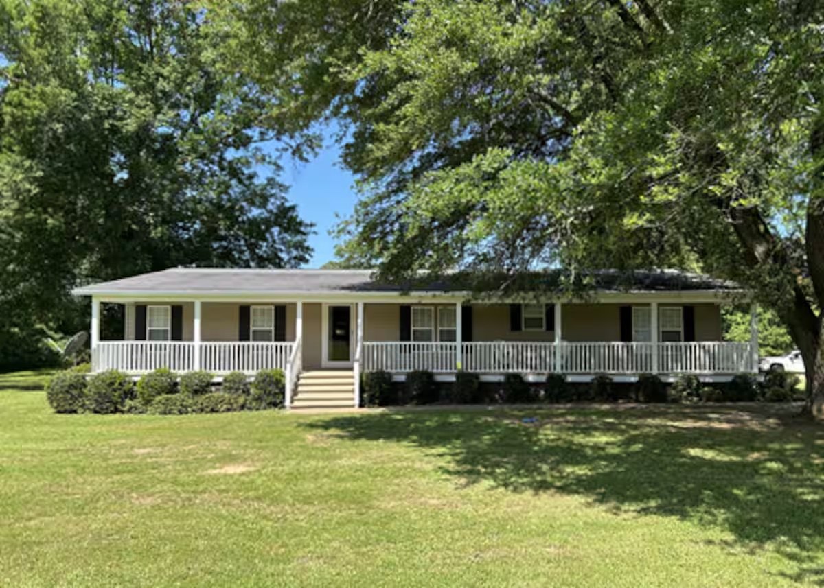 A tan ranch house with black shutters and a long white-railed front porch, shaded by large oak trees over a wide lawn