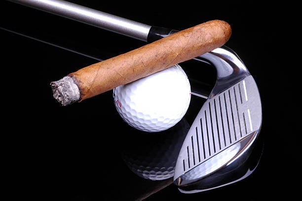 A cigar resting on a golf ball beside a golf club, on a black reflective surface
